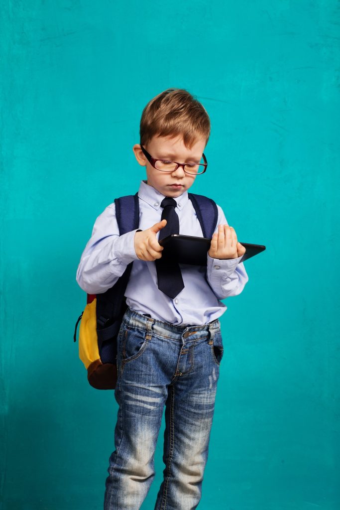 Cheerful little boy with big backpack holding digital tablet against blue background. Child playing games on a touch pad. School concept. Back to School