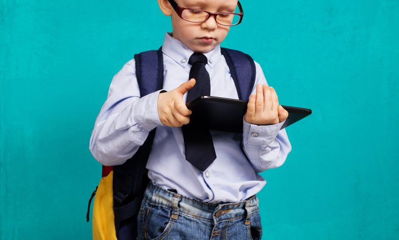 Cheerful little boy with big backpack holding digital tablet against blue background. Child playing games on a touch pad. School concept. Back to School